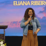 de pé, voltada ligeiramente para a esquerda do espectador, gesticulando expressivamente com a mão esquerda enquanto segura o microfone com a direita. Ela usa óculos de grau com armações redondas e finas de metal. Conduzindo a pregação "Eucaristia, Mistério celebrado no amor"