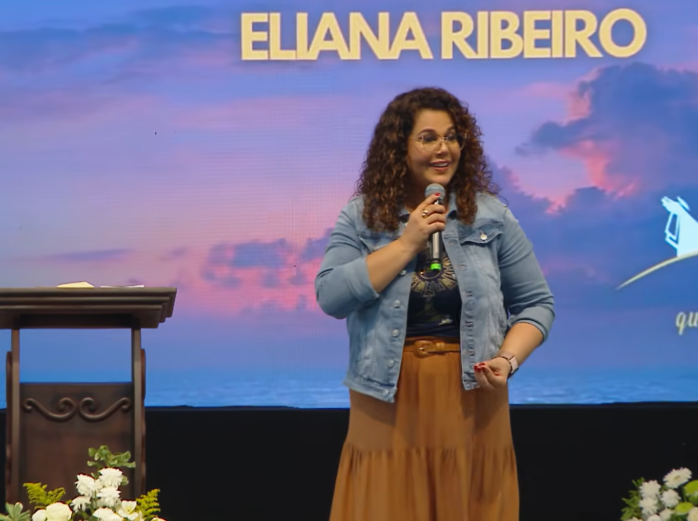 de pé, voltada ligeiramente para a esquerda do espectador, gesticulando expressivamente com a mão esquerda enquanto segura o microfone com a direita.Ela usa óculos de grau com armações redondas e finas de metal. Conduzindo a pregação "Eucaristia, Mistério celebrado no amor"