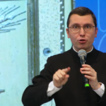 Um homem de terno preto e gola clerical fala expressivamente ao microfone, gesticulando com a mão direita. Tema: "Eucaristia, Mistério que edifica e salva"