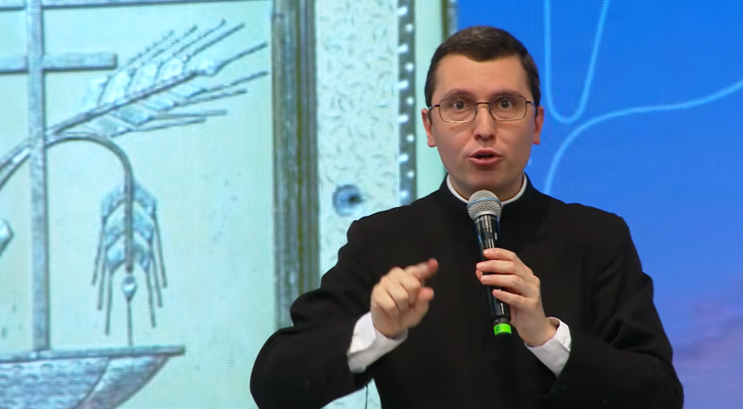 Um homem de terno preto e gola clerical fala expressivamente ao microfone, gesticulando com a mão direita. Tema: "Eucaristia, Mistério que edifica e salva"