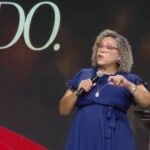 Mulher de cabelo cacheado, curto loiro usando vestido azul longo segurando com uma das mãos o microfone enquanto gesticula com a outra mão.
