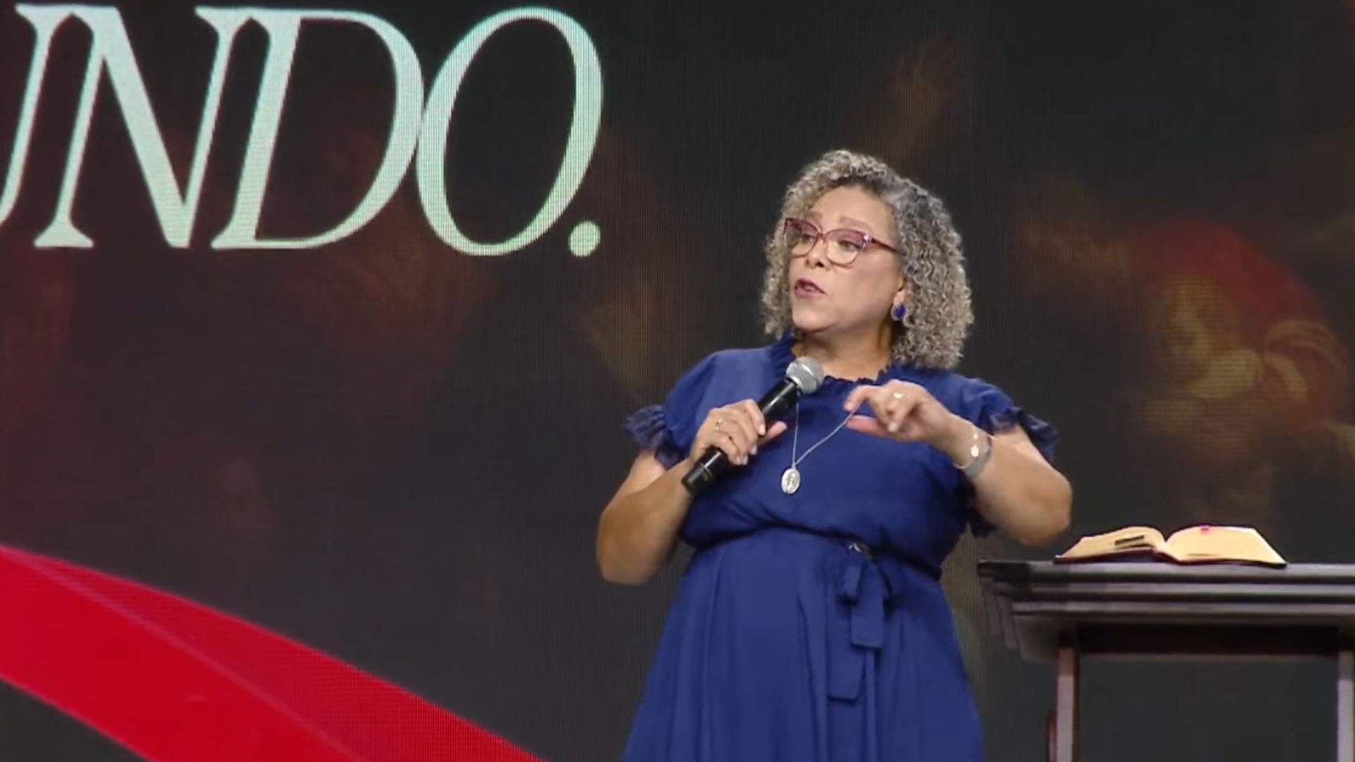 Mulher de cabelo cacheado, curto loiro usando vestido azul longo segurando com uma das mãos o microfone enquanto gesticula com a outra mão.