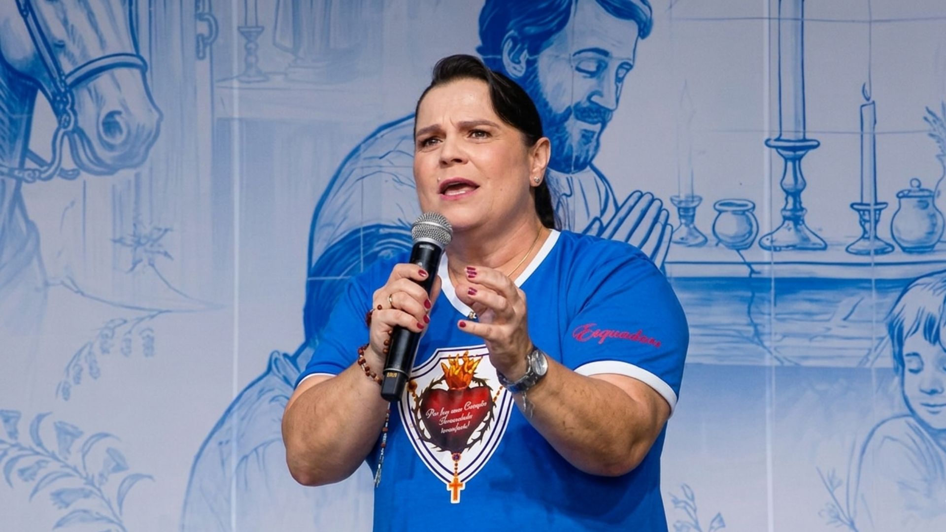 Mulher fala ao microfone em um palco.Ela veste uma camiseta azul e preta e tem cabelo preto preso. Tema " Das águas à graça Deus visita o seu povo"