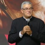 Padre de cabelo grisalho e óculos, vestindo batina preta, segurando na mão um microfone.