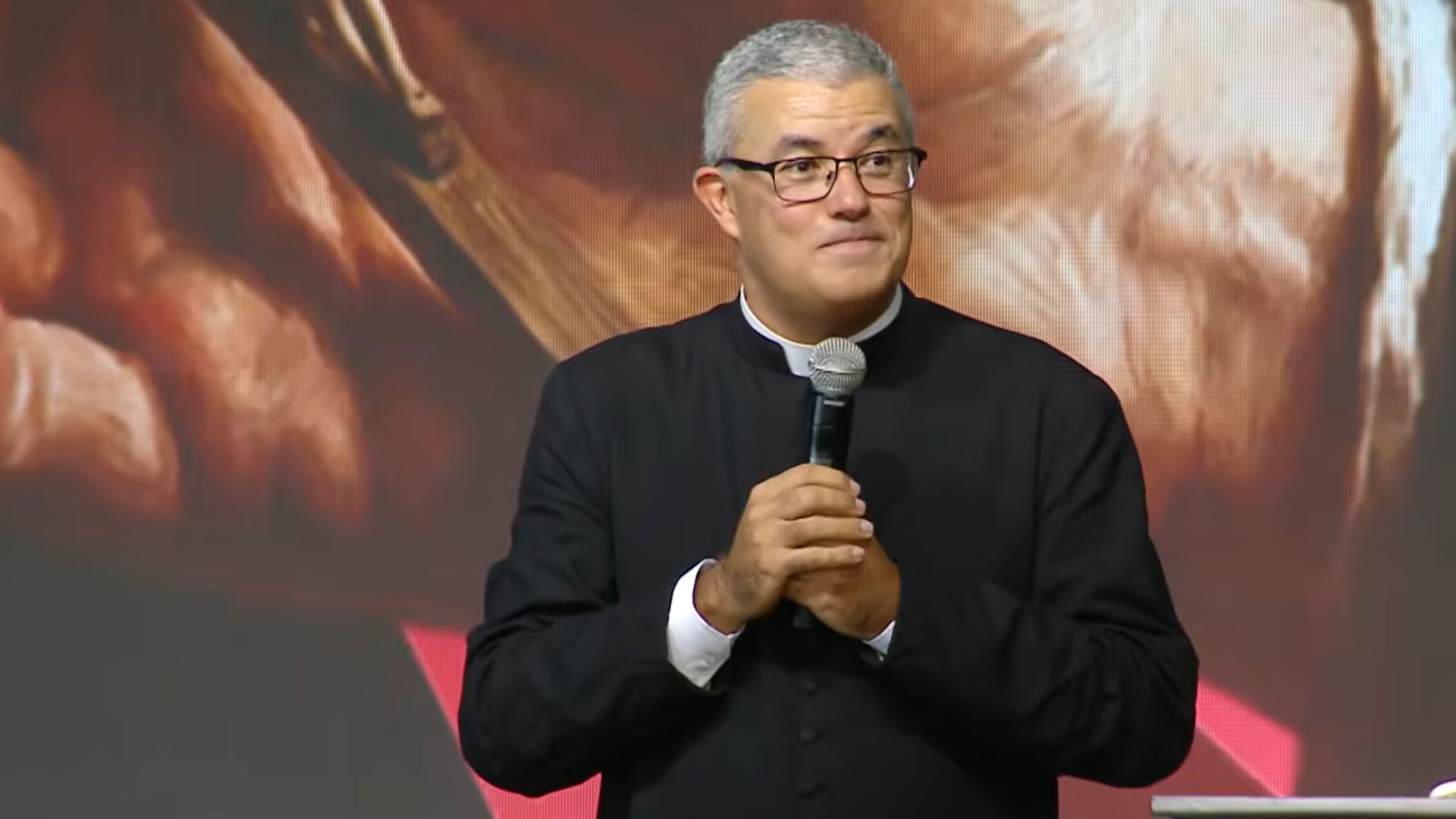 Padre de cabelo grisalho e óculos, vestindo batina preta, segurando na mão um microfone.