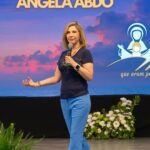 Mulher está de pé no centro de um palco, com o braço esquerdo estendido e segurando um microfone na mão direita conduzinda a pregação "Emaús, GPS divino para encontrar Jesus Ressuscitado na Eucaristia"