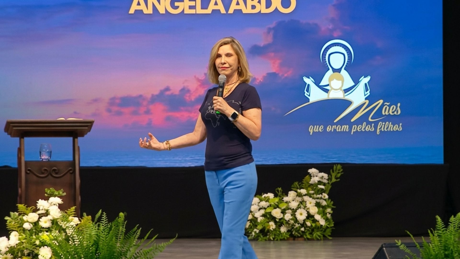 Mulher está de pé no centro de um palco, com o braço esquerdo estendido e segurando um microfone na mão direita conduzinda a pregação "Emaús, GPS divino para encontrar Jesus Ressuscitado na Eucaristia"