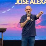 Um homem careca e de barba grisalha discursa em um palco segurando um microfone e apontando para frente. Ele está posicionado ao lado de um púlpito de madeira. Tema: "Eucaristia, Caminho seguro para a santidade"
