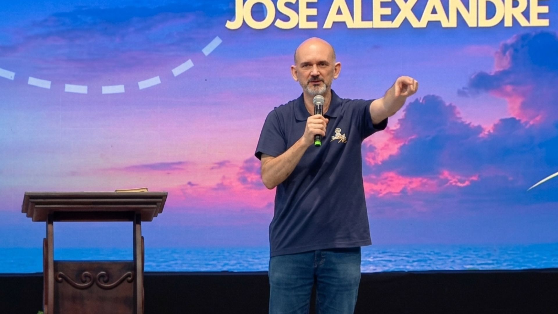 Um homem careca e de barba grisalha discursa em um palco segurando um microfone e apontando para frente. Ele está posicionado ao lado de um púlpito de madeira. Tema: "Eucaristia, Caminho seguro para a santidade"