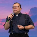 Padre usando camisa de cor azul escura com colarinho clergyman e suspens´rios pretos sobre as calças escuras e com óculos de grau. Ele segura um microfone com a mão direita e faz um gesto suave com a mão esquerda, mantendo uma expressão serena e focada enquanto conduz pregação com o tema: " Eucaristia, construindo pontes e servindo aos irmãos"