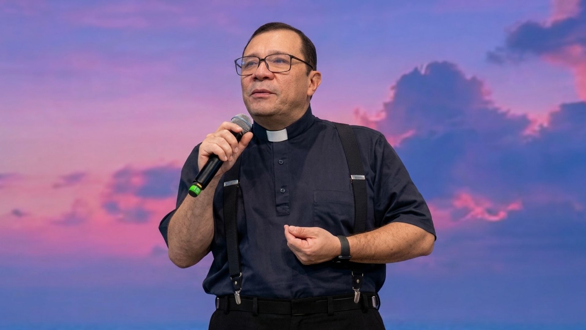 Padre usando camisa de cor azul escura com colarinho clergyman e suspens´rios pretos sobre as calças escuras e com óculos de grau. Ele segura um microfone com a mão direita e faz um gesto suave com a mão esquerda, mantendo uma expressão serena e focada enquanto conduz pregação com o tema: " Eucaristia, construindo pontes e servindo aos irmãos"