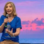 Mulher está de pé no centro de um palco, gesticulando com uma das mãos enquanto a outra segura o microfone. Tema: "Eucaristia, mistério acreditado no amor"