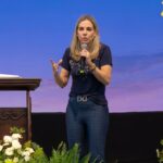 Uma mulher loira, em pé sobre um palco, falando ao microfone com a mão direita e gesticula com a mão esquerda, parecendo dar uma pregação com o tema "Eucaristia, presente de Deus para o mundo"