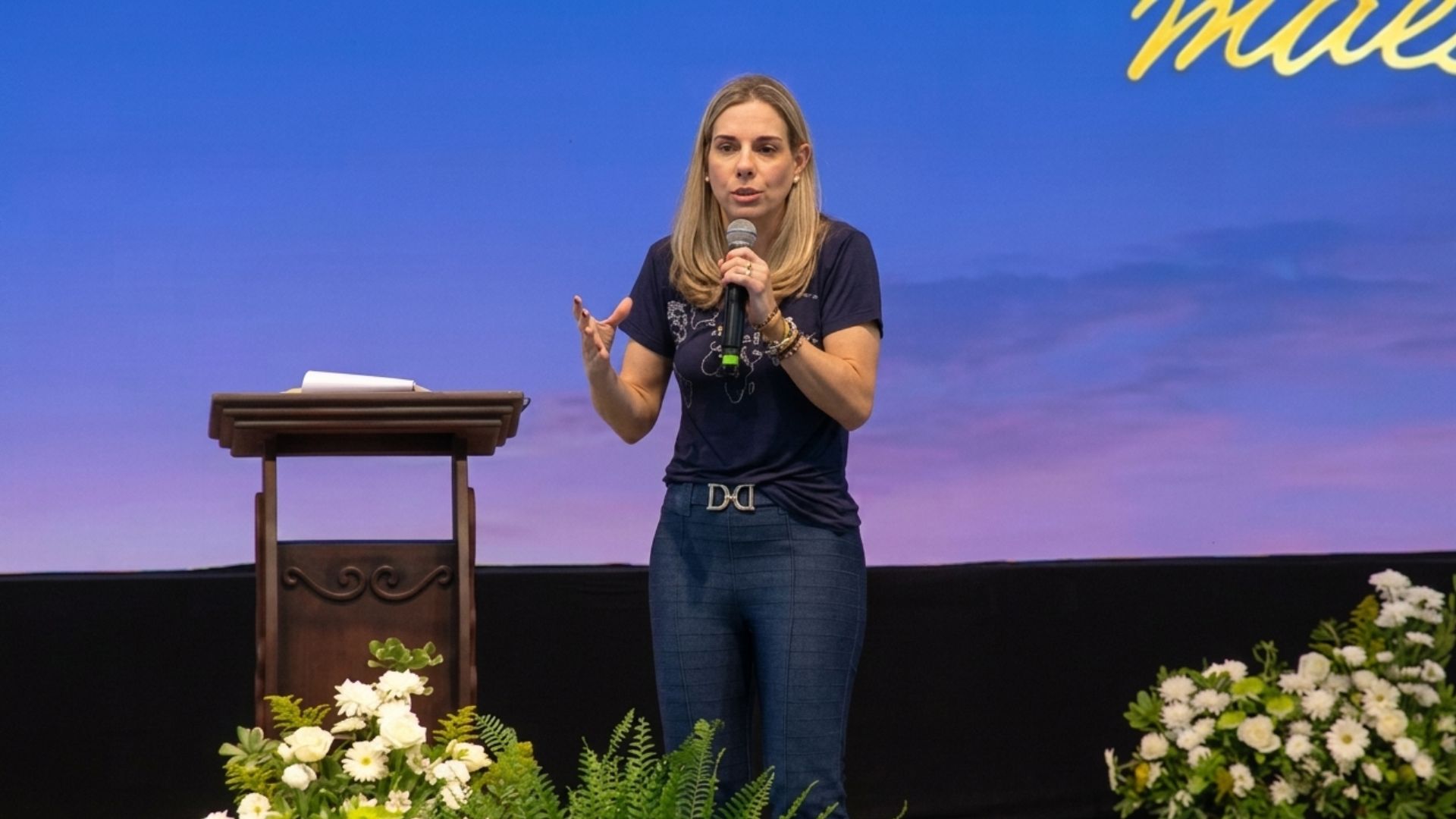 Uma mulher loira, em pé sobre um palco, falando ao microfone com a mão direita e gesticula com a mão esquerda, parecendo dar uma pregação com o tema "Eucaristia, presente de Deus para o mundo"