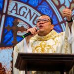 Um padre com vestimentas brancas fala ao microfone posicionado atrás de um púlpito de madeira. O fundo apresenta um painel artístico colorido em estilo vitral com símbolos religiosos e uma cruz. Tema: "Como é grande, ó Pai, a Vossa Misericórdia!"