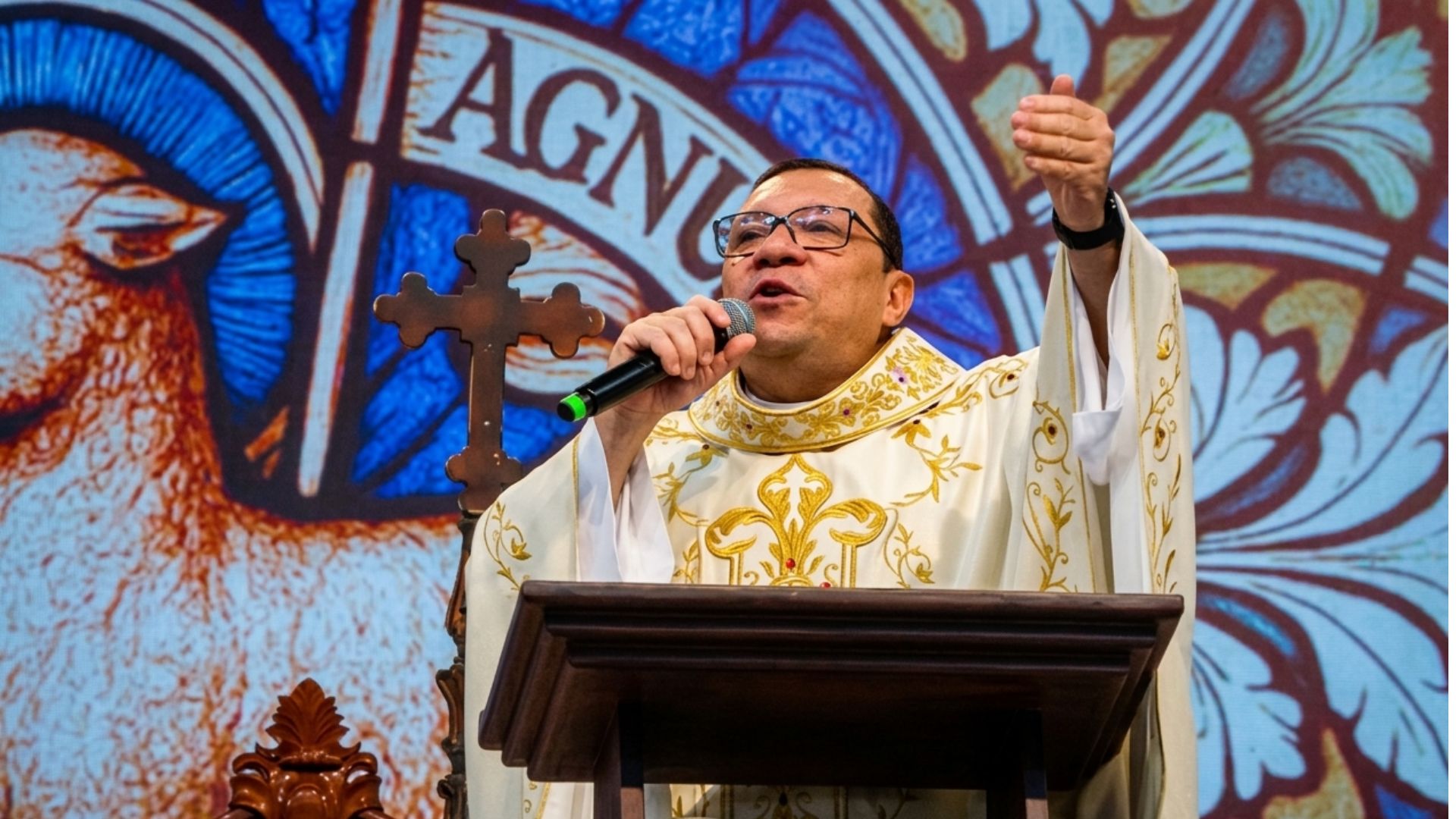 Um padre com vestimentas brancas fala ao microfone posicionado atrás de um púlpito de madeira.O fundo apresenta um painel artístico colorido em estilo vitral com símbolos religiosos e uma cruz. Tema: "Como é grande, ó Pai, a Vossa Misericórdia!"