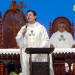 Padre discursa com um microfone em um altar durante uma celebração religiosa. Ele veste túnica branca e está acompanhado por outros religiosos sentados ao fundo. Tema: "