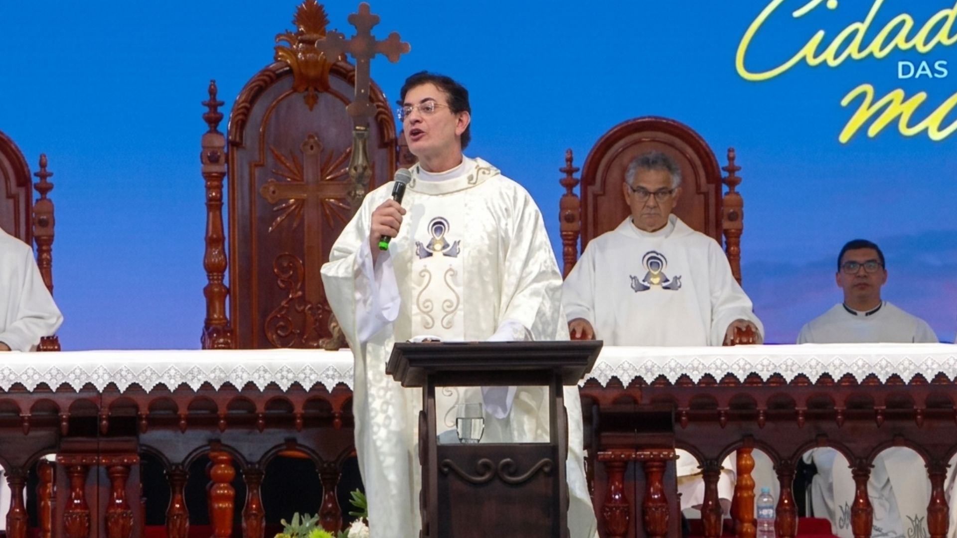 Padre discursa com um microfone em um altar durante uma celebração religiosa.Ele veste túnica branca e está acompanhado por outros religiosos sentados ao fundo. Tema: "Mães Eucarísticas reavivando a fé no encontro com Cristo"