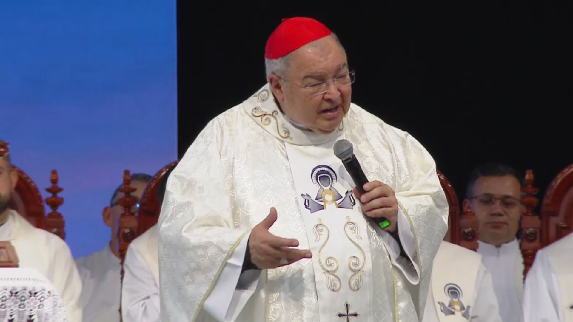 Dom Orani Tempesta, vestindo paramentos litúrgicos brancos e solidéu vermelho, fala ao microfone e gesticula durante uma celebração religiosa. Tema "Jesus, o Pão da Vida"