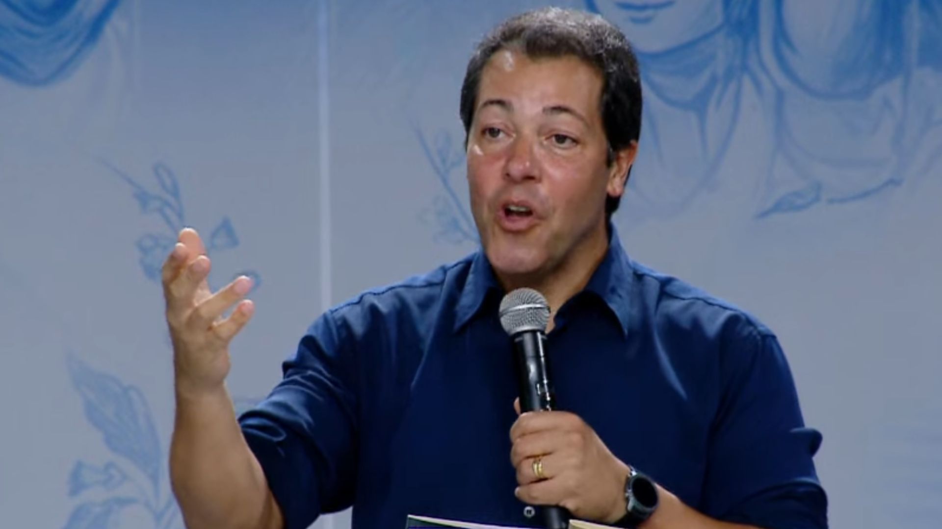 Homem segurando o microfone enquanto gesticula com uma das mãos. Tema: "Maria, a Mãe que intercede: Dai-nos a bênção!"