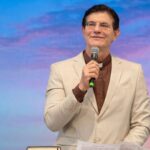 Sacerdote com gola clerical sorrindo, de óculos, terno bege e camisa marrom, segurando um microfone enquanto fala em um púlpito com um livro aberto. Tema: "Na escola de Maria, ser Mãe Eucarística"