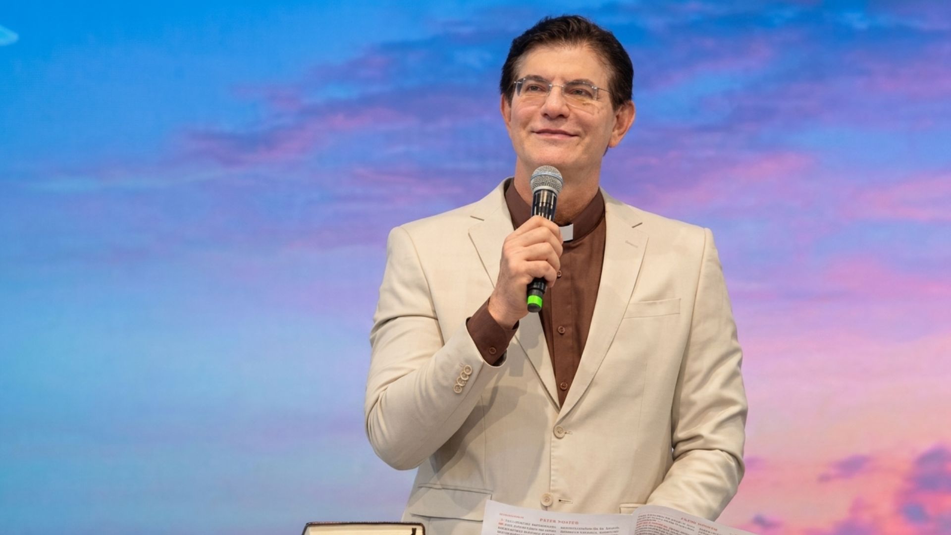 Sacerdote com gola clerical sorrindo, de óculos, terno bege e camisa marrom, segurando um microfone enquanto fala em um púlpito com um livro aberto. Tema: "Na escola de Maria, ser Mãe Eucarística"