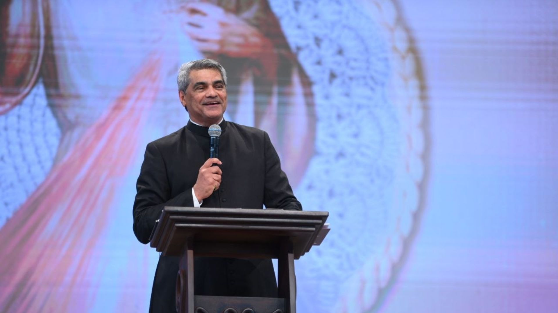 Padre de cabelos grisalhos e vestindo uma batina preta atras de um púlíto enquanto fala segurando um microfone. Ao fundo imagem de Jesus Misericordioso. Ele conduz a pregação com o tema "Nada me impedirá de conceder graças"