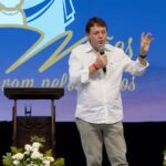 Homem usando camisa branca de botões e calça clara, segurando uma das mãos com o microfone enuanto gesticula com a outra. Tema: "O Milagre da Eucaristia e o testemunho da Serva de Deus Lola"