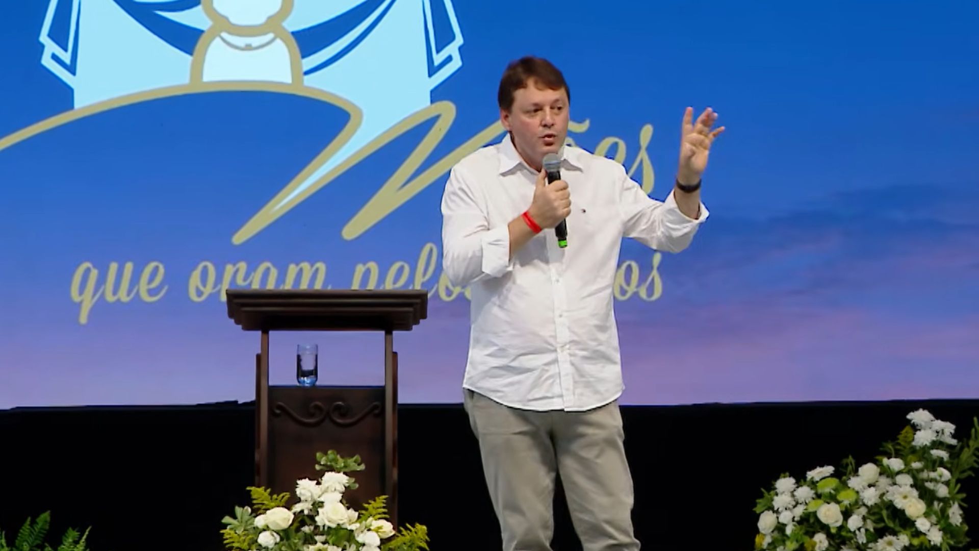 Homem usando camisa branca de botões e calça clara, segurando uma das mãos com o microfone enuanto gesticula com a outra. Tema: "O Milagre da Eucaristia e o testemunho da Serva de Deus Lola"