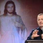Pregador usando roupa preta gesticulando com uma das mãos e segurando o microfone com a outra. Ao fundo imagem de Jesus misericordioso simbolizando a Festa da Misericórdia.