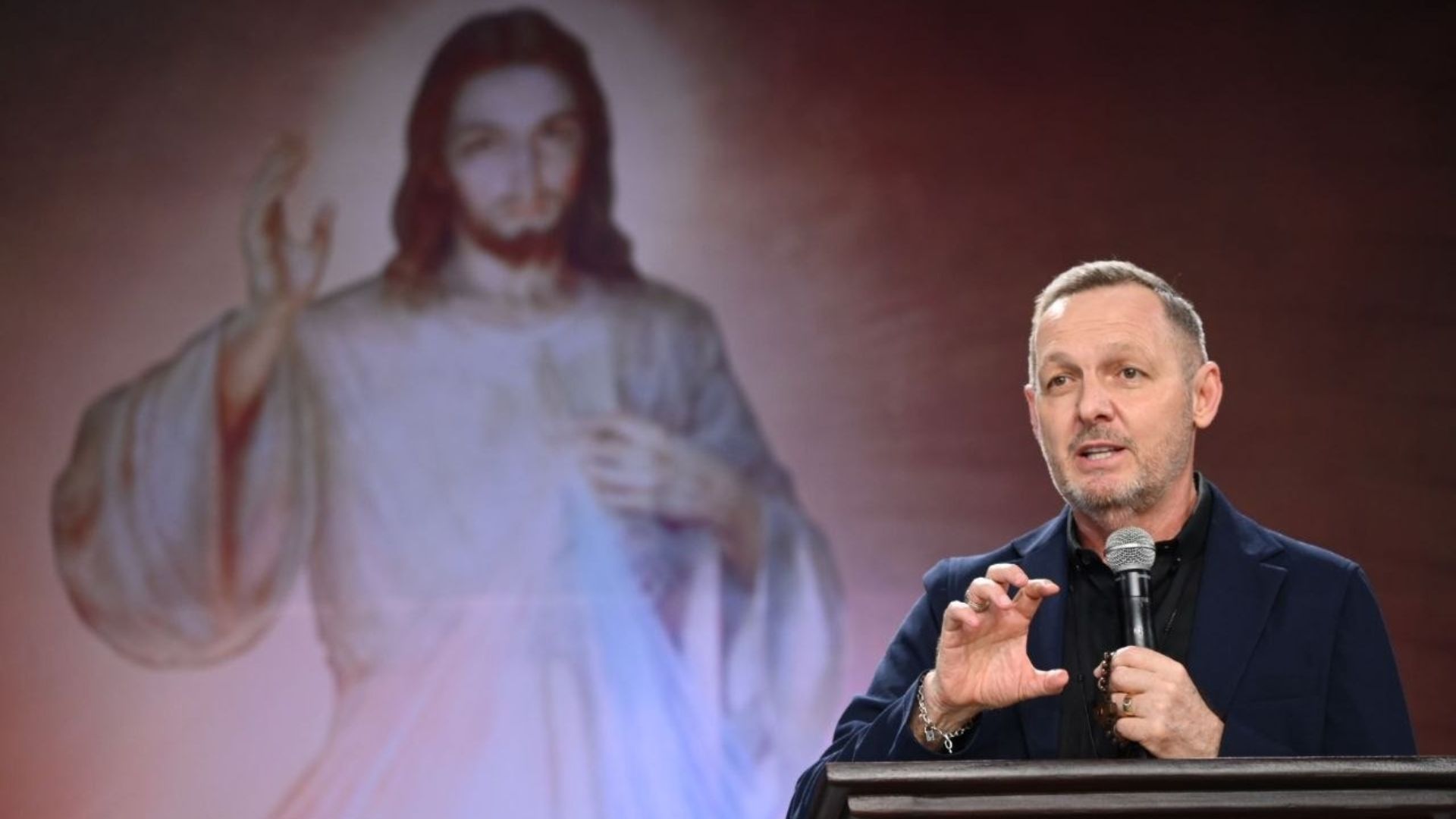 Pregador usando roupa preta gesticulando com uma das mãos e segurando o microfone com a outra. Ao fundo imagem de Jesus misericordioso simbolizando a Festa da Misericórdia.