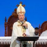 Um bispo católico de mitra e vestes brancas discursa ao microfone em um altar durante uma celebração religiosa conduzindo a homilia sobre "O poder da oração das mães e a Eucaristia"