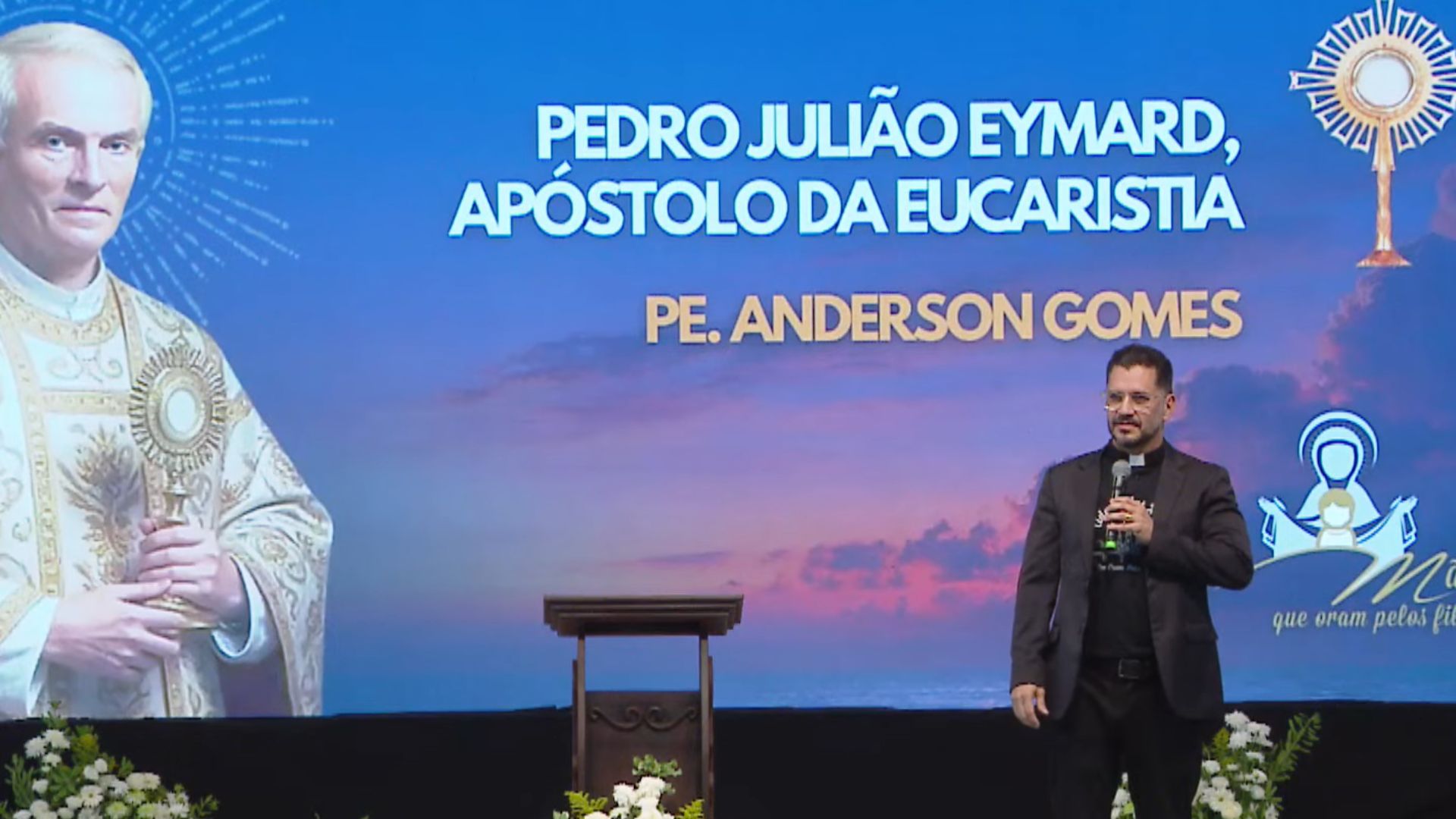 Sacerdote ministra uma palestra em um palco, posicionado à frente de um grande telão com o título "Pedro Julião Eymard, Apóstolo da Eucaristia".