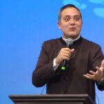 Um homem de terno preto e gola clerical fala expressivamente ao microfone, gesticulando com a mão. Tema: "Eucaristia, fonte de serviço aos irmãos".