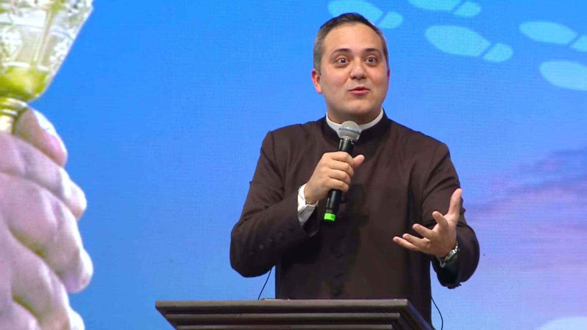 Um homem de terno preto e gola clerical fala expressivamente ao microfone, gesticulando com a mão. Tema: "Eucaristia, fonte de serviço aos irmãos".