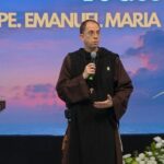 Sacerdote usando hábito religioso marrom, palestrando ao microfone em um palco com púlpito, arranjos de flores e um telão azul ao fundo. Tema: "Presença real e adoração Eucarística segundo a tradição e CIC"