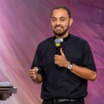 Um padre jovem, com barba e vestes clericais pretas, sorri enquanto segura um microfone e gesticula com a outra mão. Ele está em um palco, posicionado à frente de um fundo com tons de roxo e luzes suaves. À esquerda, vê-se parte de um púlpito.