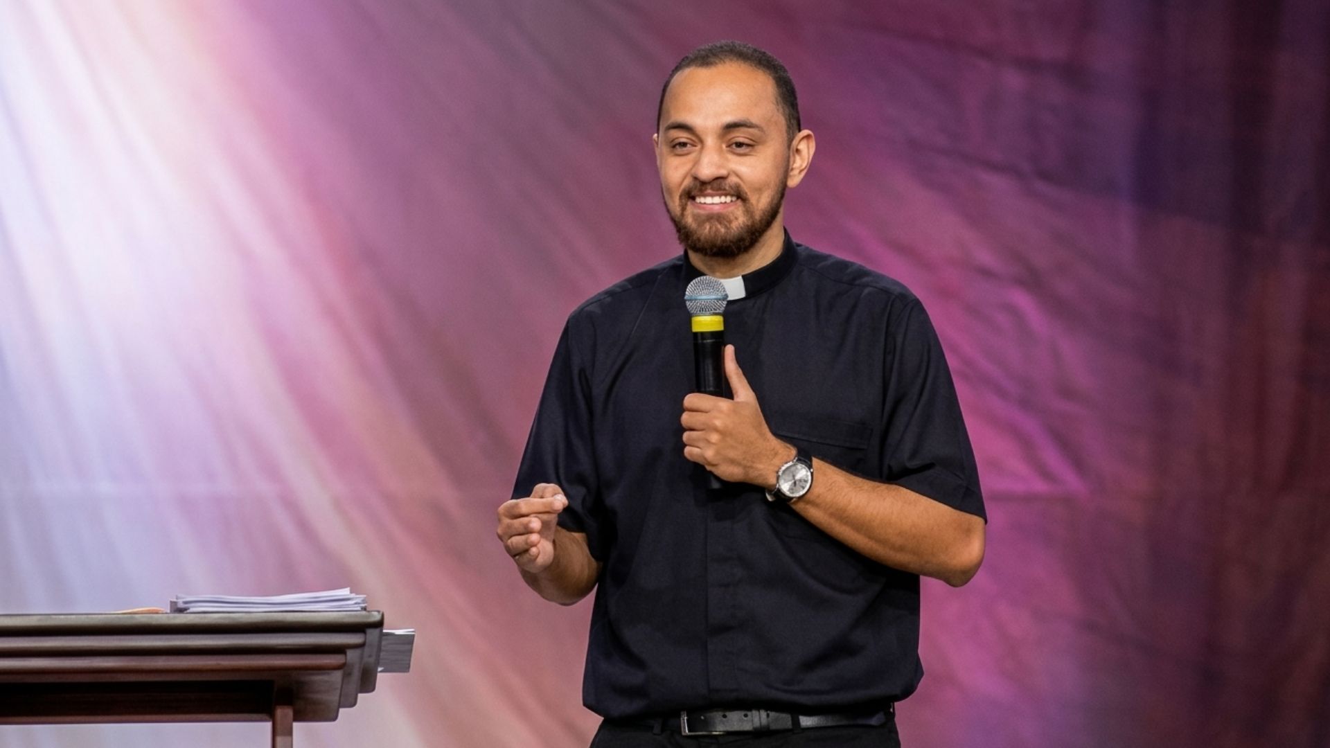 Um padre jovem, com barba e vestes clericais pretas, sorri enquanto segura um microfone e gesticula com a outra mão. Ele está em um palco, posicionado à frente de um fundo com tons de roxo e luzes suaves. À esquerda, vê-se parte de um púlpito.