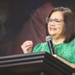 Mulher com camiseta verde e microfone na mão num pulpito para realizar pregação