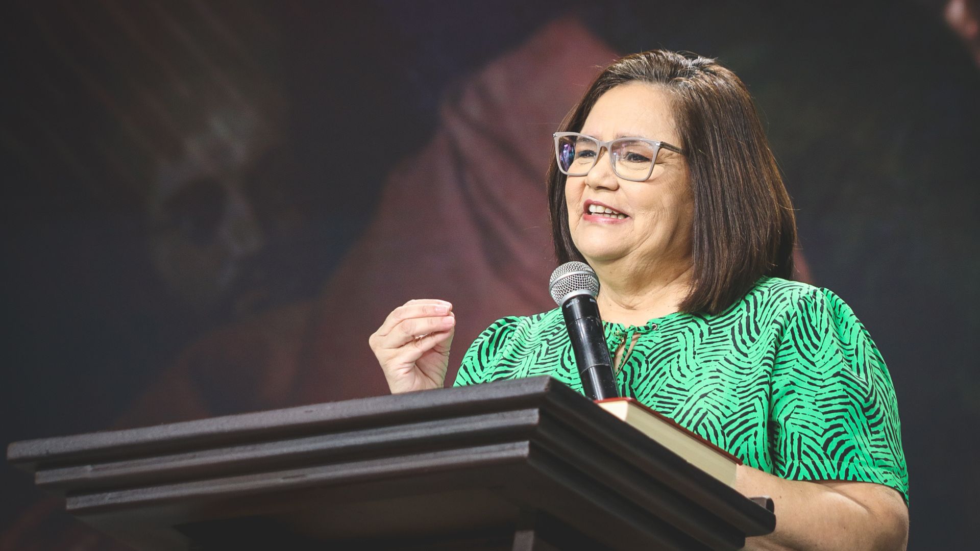 Mulher com camiseta verde e microfone na mão num pulpito para realizar pregação