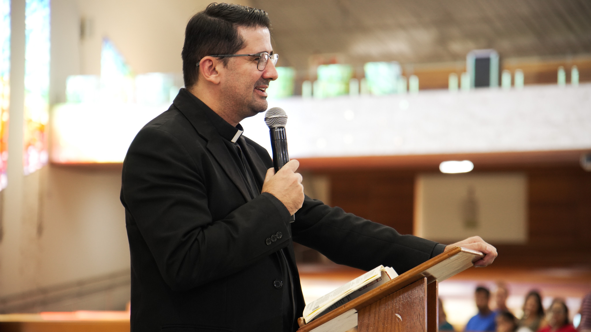 O padre de óculos e vestes pretas prega com um microfone. Tema: "O mistério da filiação Divina e o caminho da misericórdia"