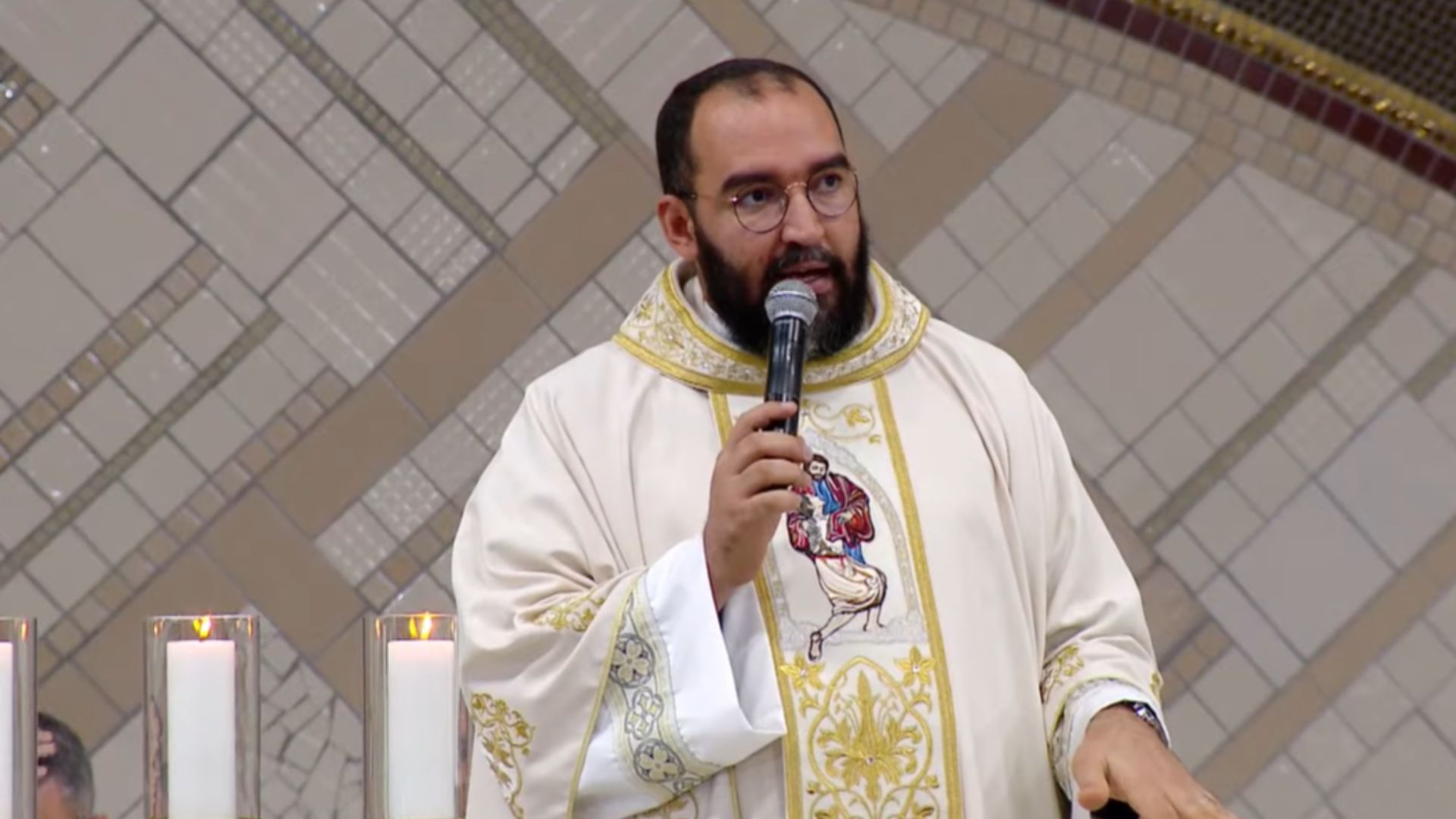 Um padre de barba e óculos, vestindo uma túnica litúrgica branca com detalhes dourados, fala ao microfone durante uma homilia. Tema: "Jesus Cristo: o único Caminho, a Verdade e a Vida"