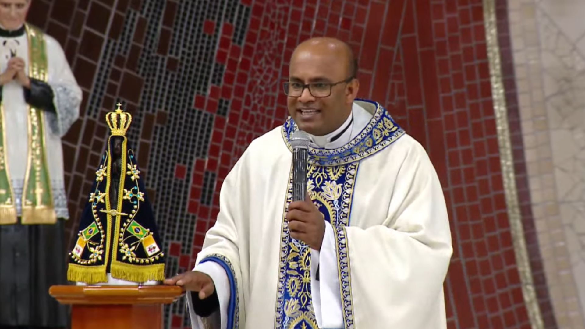 Um sacerdote com óculos e vestes brancas e azuis segura um microfone e gesticula ao lado da imagem de Nossa Senhora Aparecida. Tema: "Nossa Senhora intercede pela restauração da nossa vida"