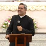 Padre vestindo batina preta e colarinho clerical, fala ao microfone posicionado atrás de um ambão de madeira. Ao fundo, avista-se o altar ornamentado com uma toalha litúrgica branca e arranjos de flores delicadas.