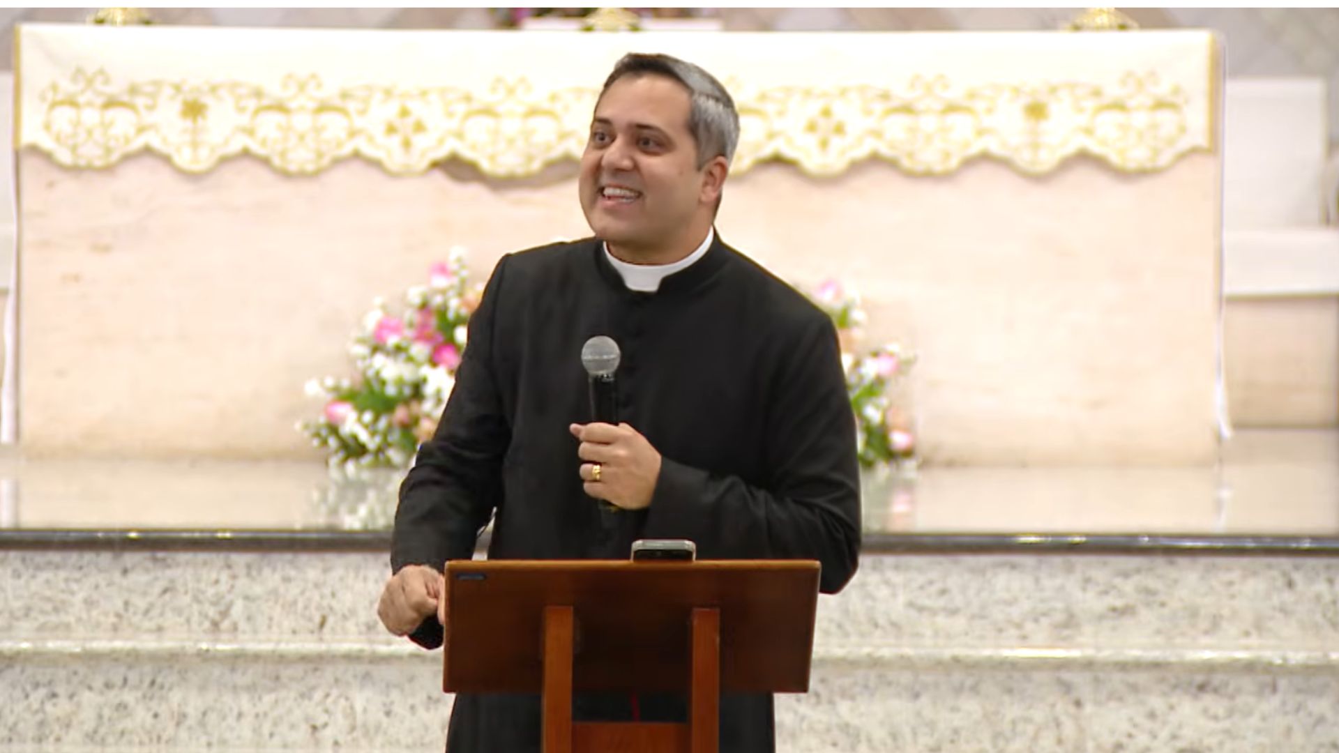 Padre vestindo batina preta e colarinho clerical, fala ao microfone posicionado atrás de um ambão de madeira.Ao fundo, avista-se o altar ornamentado com uma toalha litúrgica branca e arranjos de flores delicadas.