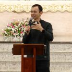 Padre jovem, sorridente e de batina preta, prega com entusiasmo ao microfone num ambão de madeira. O fundo mostra o altar da igreja com um pano dourado e um belo arranjo de flores rosa e brancas.