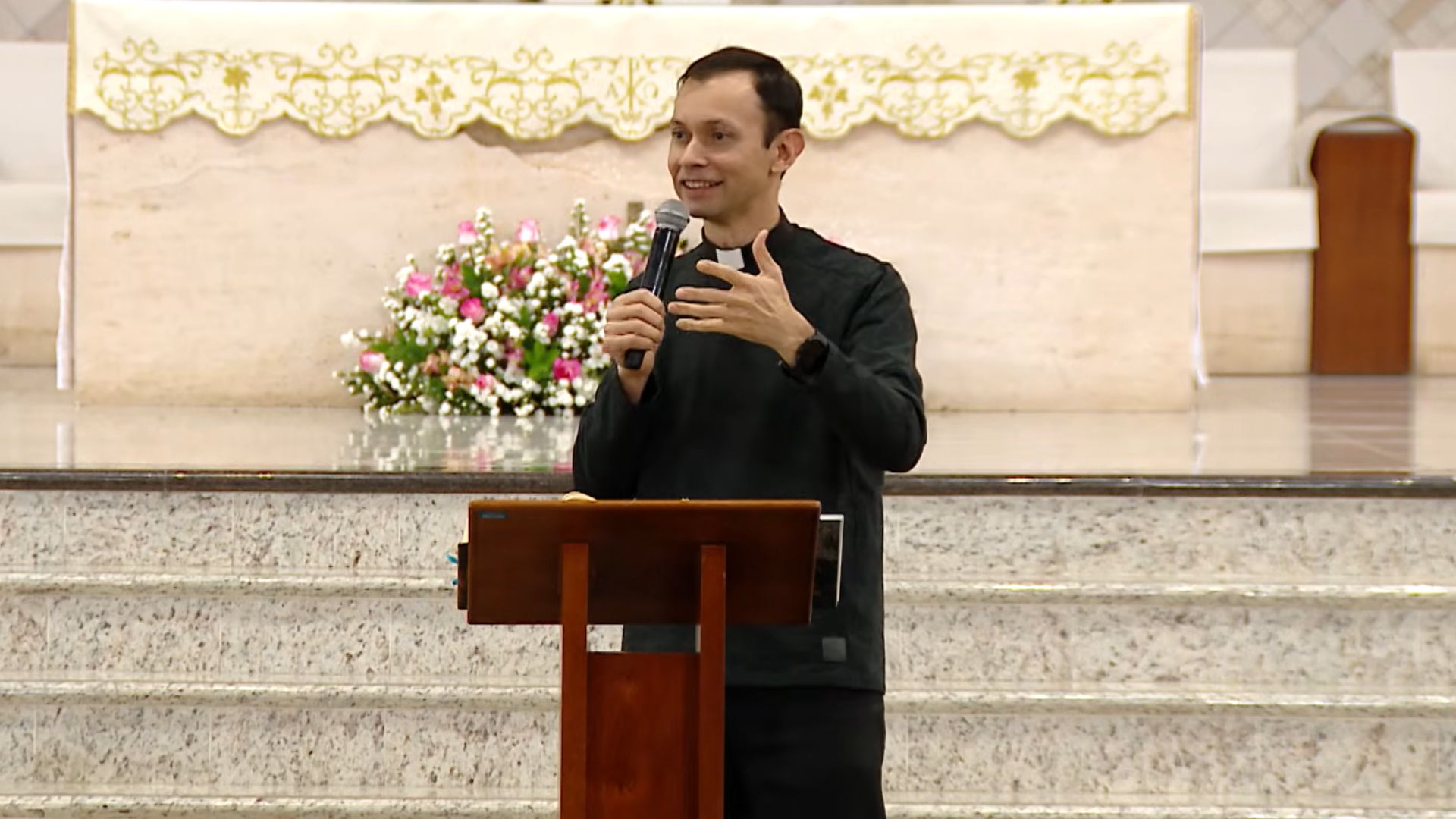 Padre jovem, sorridente e de batina preta, prega com entusiasmo ao microfone num ambão de madeira.O fundo mostra o altar da igreja com um pano dourado e um belo arranjo de flores rosa e brancas.