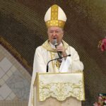 Bispo com vestes brancas e mitra dourada fala ao microfone em um ambão decorado com o símbolo Alfa e Ômega. Ao fundo, destaca-se uma parede em mosaico e, à direita, uma imagem de Nossa Senhora Auxiliadora com o Menino Jesus. Tema: "O amor incondicional de Deus em nossa vida"