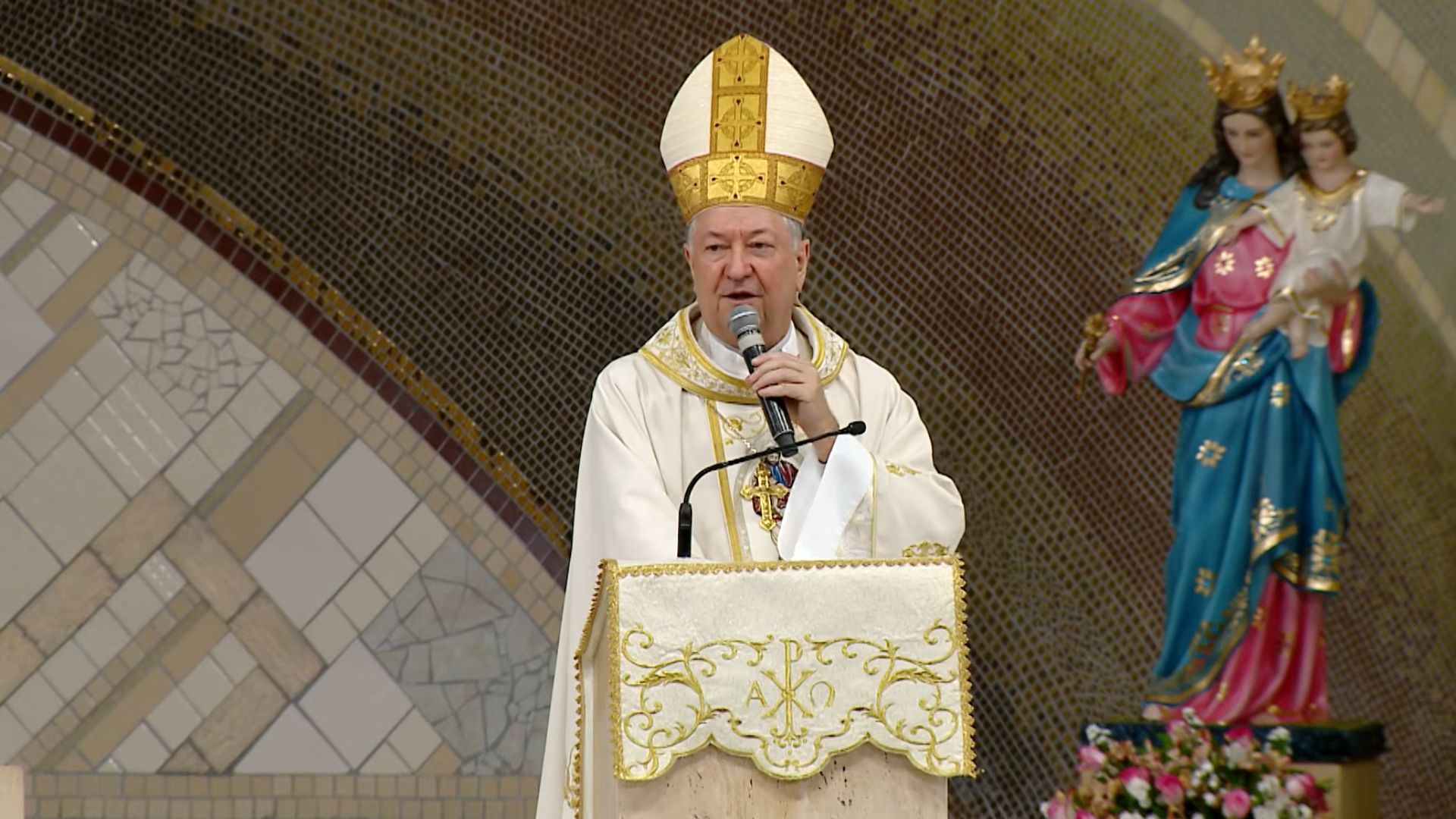 Bispo com vestes brancas e mitra dourada fala ao microfone em um ambão decorado com o símbolo Alfa e Ômega.Ao fundo, destaca-se uma parede em mosaico e, à direita, uma imagem de Nossa Senhora Auxiliadora com o Menino Jesus.