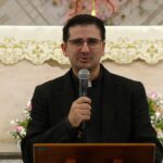 O padre de óculos e vestes pretas prega com um microfone no altar da igreja. Atrás dele, há um detalhado arranjo de flores e uma toalha litúrgica bordada. Tema: "O mistério da filiação Divina e o caminho da misericórdia"